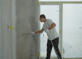 A man uses a spray gun to paint an interior wall in a room under renovation.